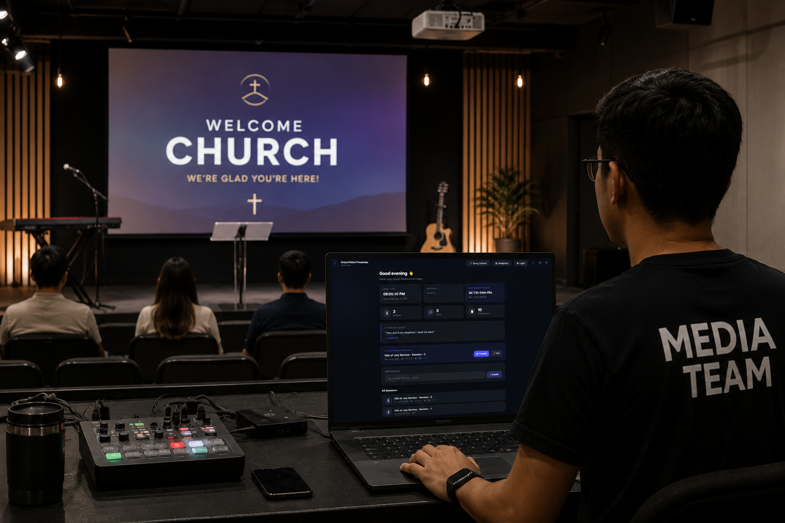 Close-up of hands running GraceVision Presenter on a tablet and laptop from the sound booth, with a worshipping congregation and lyrics on the big screen behind them.